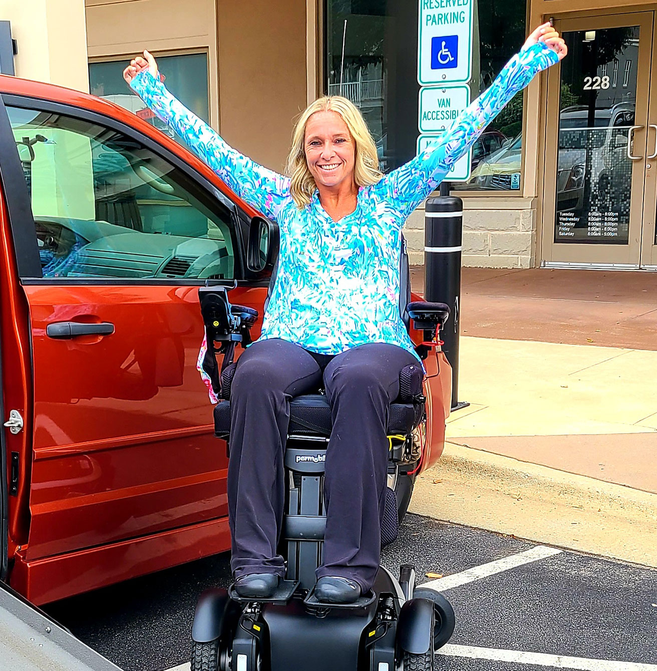 Ali in her wheelchair dressed in a blue and white shirt by a red van in a victory pose.