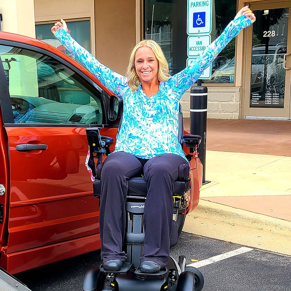 Ali with her arms up by a Orange adapted van with wheelchair lifted up An elevated position